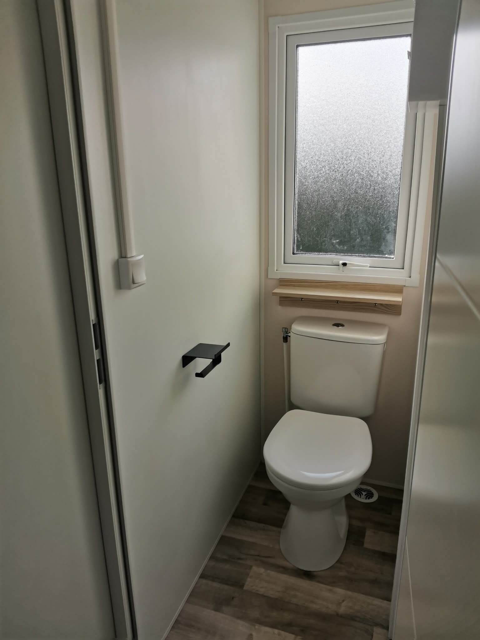 Un baño pequeño y compacto con inodoro blanco, una ventana sobre el inodoro y piso de madera.