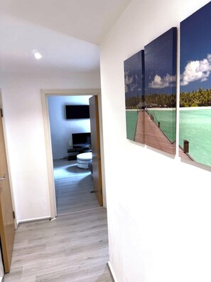 A hallway with a wooden floor leading to a bathroom with a toilet and a sink. There are two framed pictures on the wall depicting a tropical beach scene. - Charming 1 bedroom flat in London Excel Docklands (London)