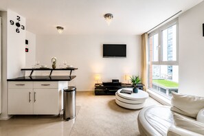 A modern living room with a sofa, a TV, a dining area with a table and chairs, and a kitchen with cabinets and a trash bin. - Charming 1 bedroom flat in London Excel Docklands (London)