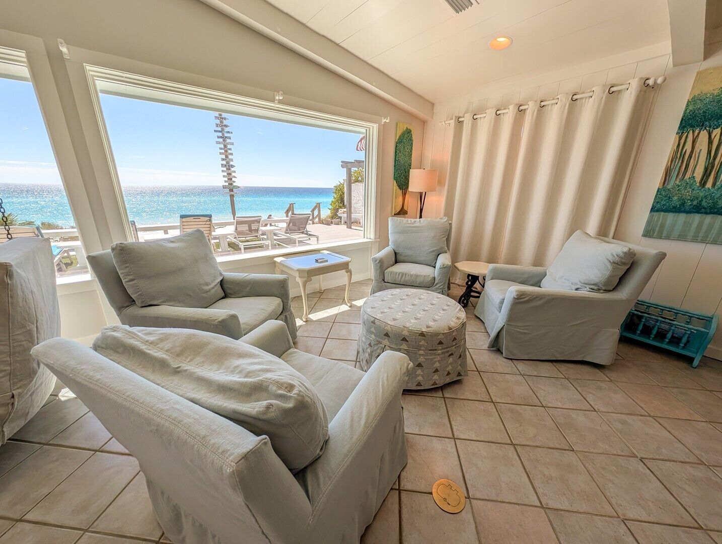 A sunlit living room with a view of the sea, featuring a sofa, armchairs, a small table, and a lamp.