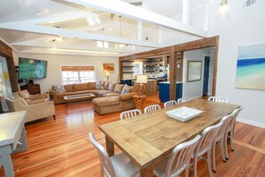 A spacious living area with a wooden dining table, multiple seating options, and a kitchen in the background. - Luxury Old Seagrove Retreat, Walk to Beach / Seaside, Private Heated Pool, Game Room, Pet Friendly (Seagrove Beach)
