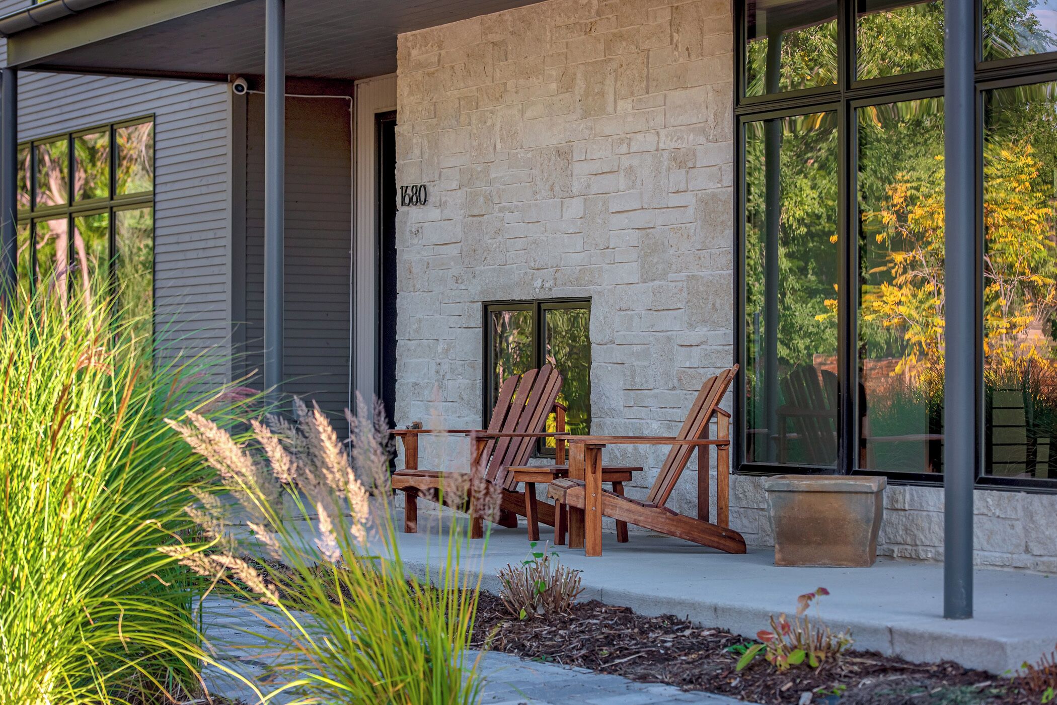 A modern house with a stone facade, two wooden chairs outside, and a large window reflecting trees.