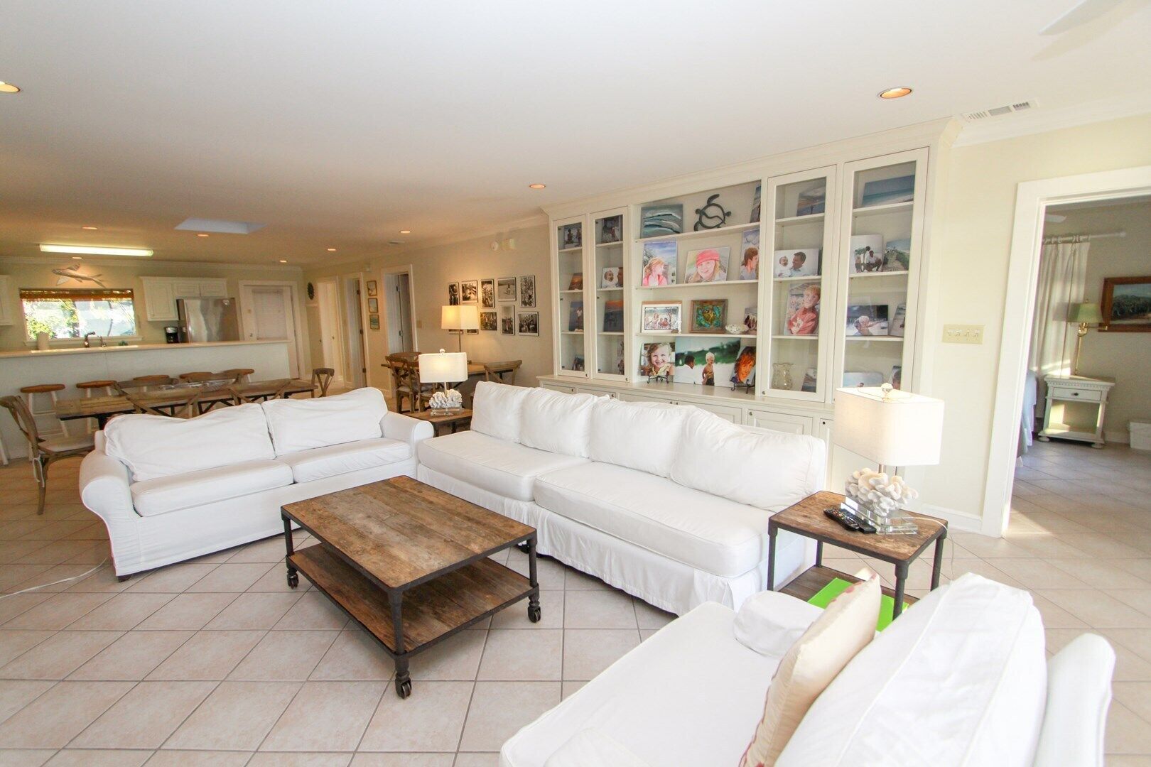 Ein geräumiges Wohnzimmer mit einem weißen modularen Sofa, einem Holztisch und einem Esstisch mit Glasplatte. Im Hintergrund ist eine Einbau-Bücherreol mit diversen Gegenständen und eine Küchenzeile zu sehen.