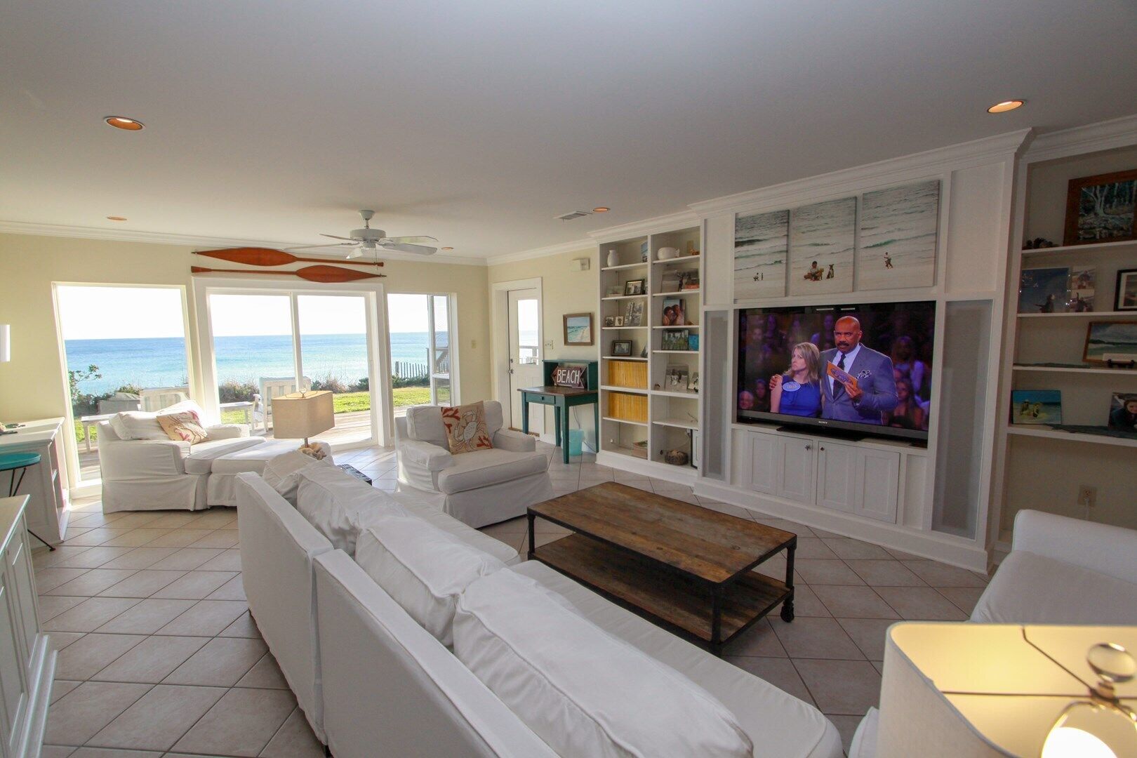 A spacious living room with a large TV, white furniture, and a view of the ocean.