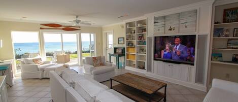 A spacious living room with a large TV, white furniture, and a view of the ocean.