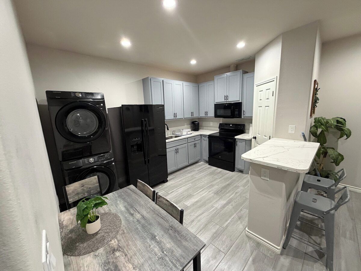 Una cocina funcional con lavadora, refrigerador, microondas y un pequeño comedor con mesa y sillas.