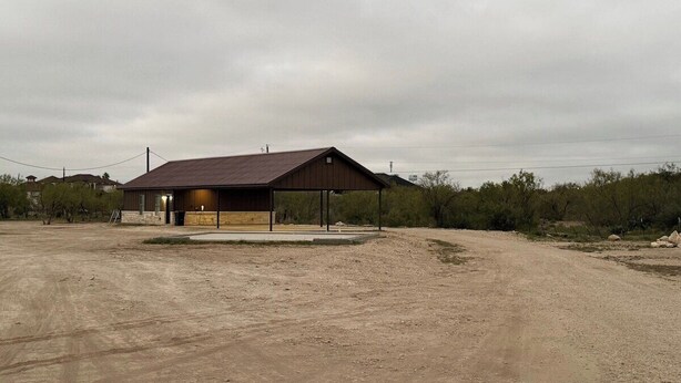 A building with a covered outdoor area, a dirt ground, and sparse vegetation in the background.