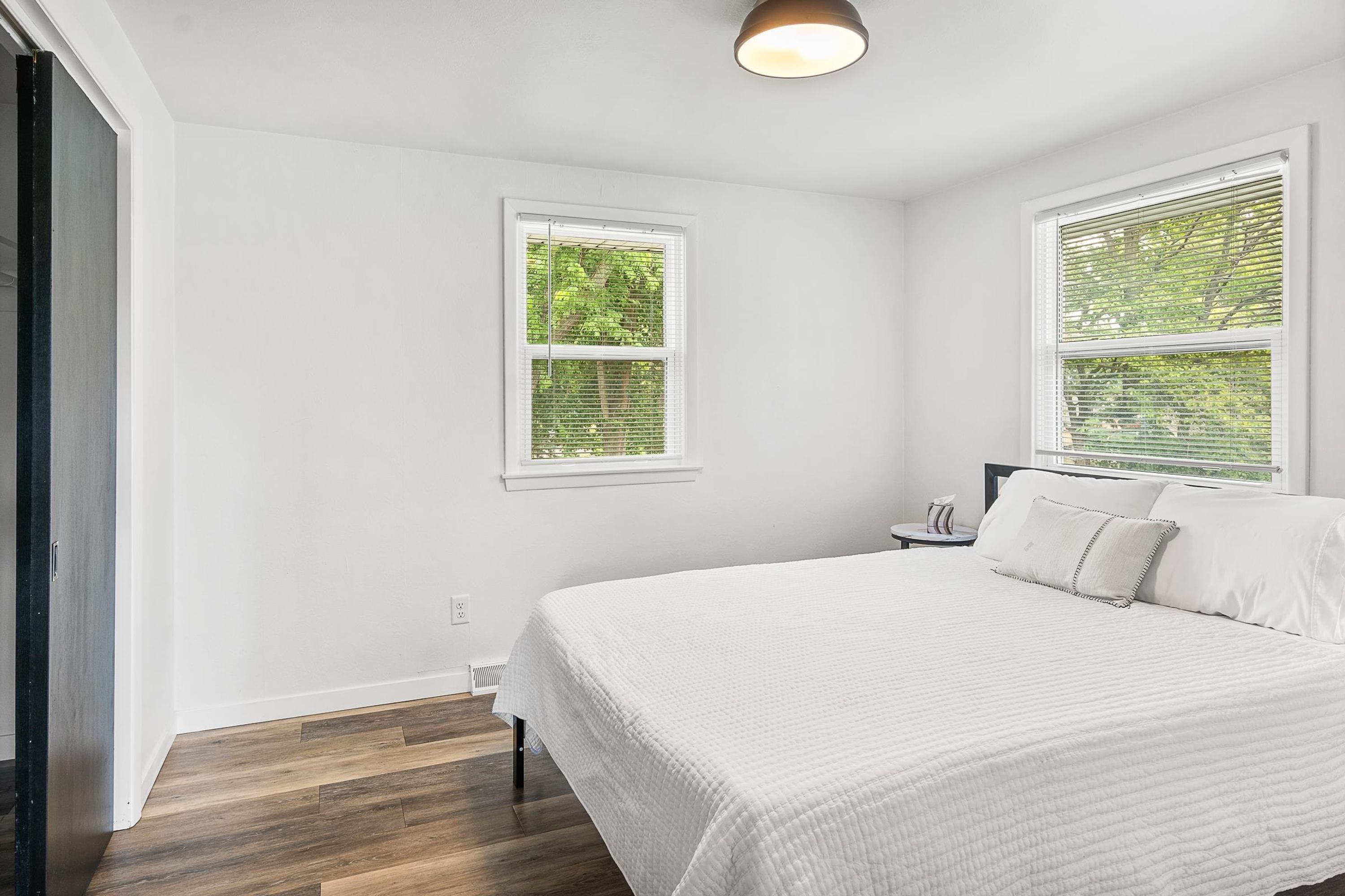 Une chambre à coucher avec un lit, deux fenêtres, une table de chevet et une prise électrique au mur.