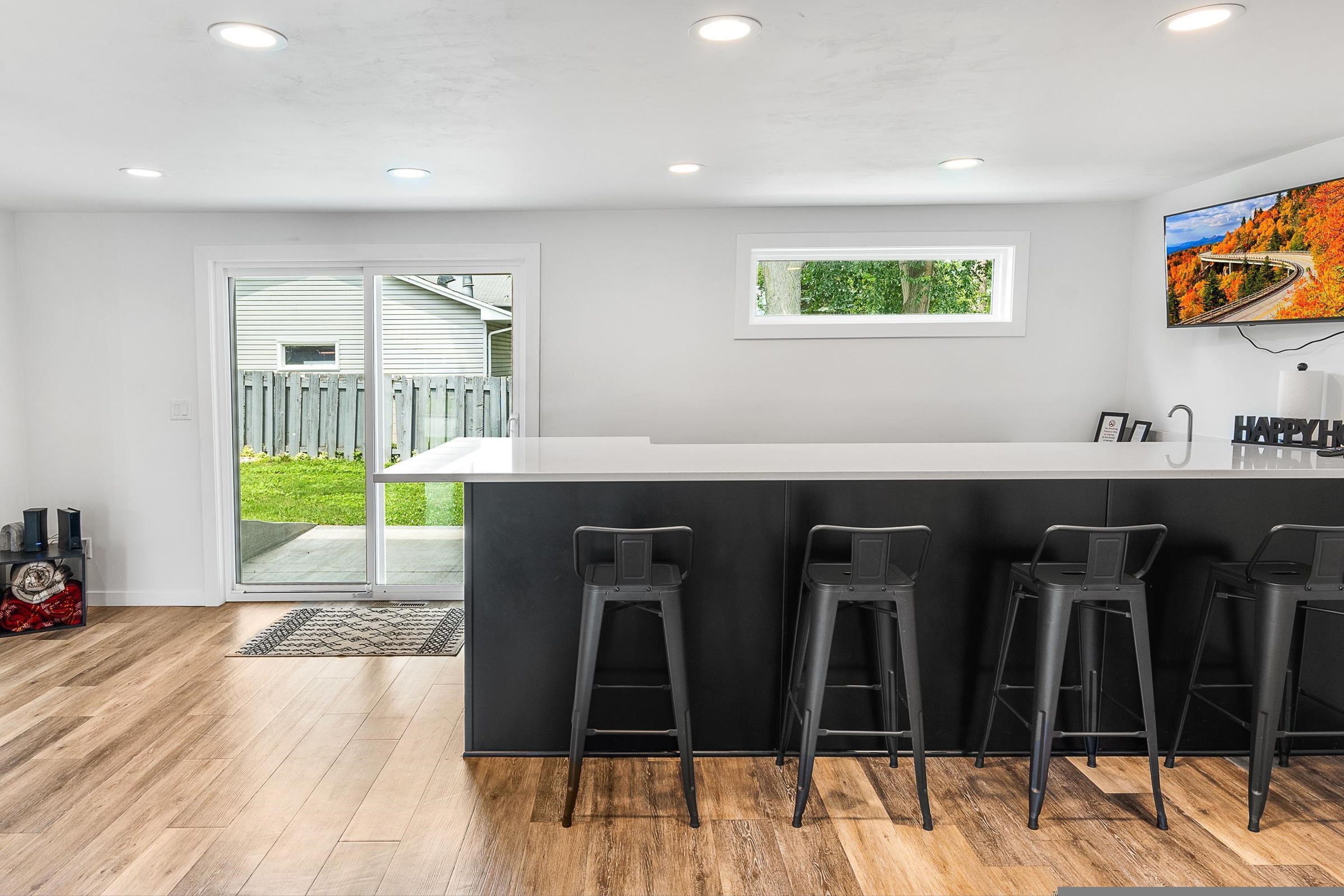 A modern kitchen with a central island, bar stools, recessed ceiling lights, and a view of the outdoors.