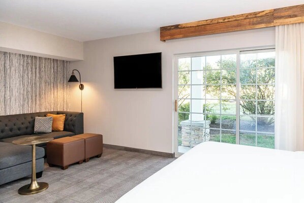 Een hotelkamer met een bank, poufjes, een klein rond tafeltje, een flatscreen televisie en een groot raam met uitzicht op groen.