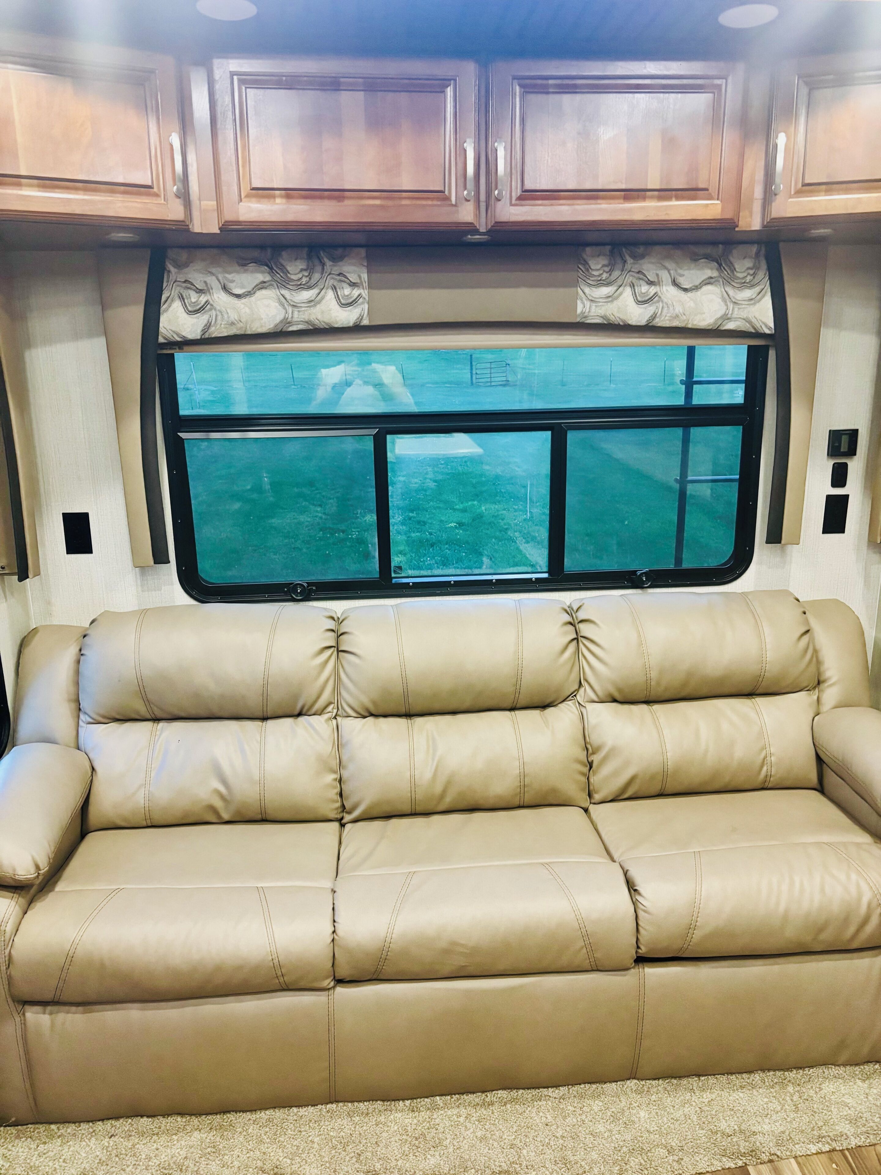 A tan leather sofa in a RV interior with wooden cabinets above and a window with a view of a grassy area.