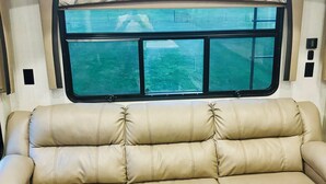 Un canapé en cuir marron dans l’intérieur d’un camping-car, avec des placards en bois au-dessus et une fenêtre donnant sur un espace verdoyant.