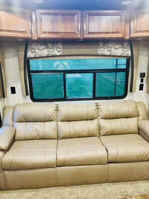 A tan leather sofa in a RV interior with wooden cabinets above and a window with a view of a grassy area. - Cozy All Season 5th wheel camper in tranquil Greenup, ky. (Greenup)