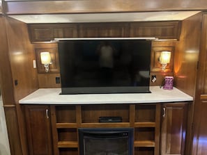 A kitchen with a large black flat-screen TV, wooden cabinets, and two wall-mounted lamps. - Cozy All Season 5th wheel camper in tranquil Greenup, ky. (Greenup)