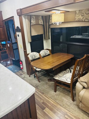 A dining area with a wooden table and chairs, a fire extinguisher, and a kitchen area with a stove and cabinets. - Cozy All Season 5th wheel camper in tranquil Greenup, ky. (Greenup)