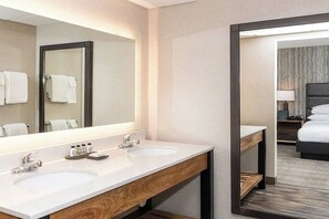 Een hotelbadkamer met twee wastafels, een grote spiegel en uitzicht op een slaapkamer met een bed en nachtkastje.