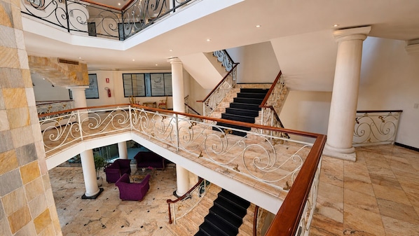 A grand staircase with ornate railings, marble flooring, and a seating area with purple armchairs.