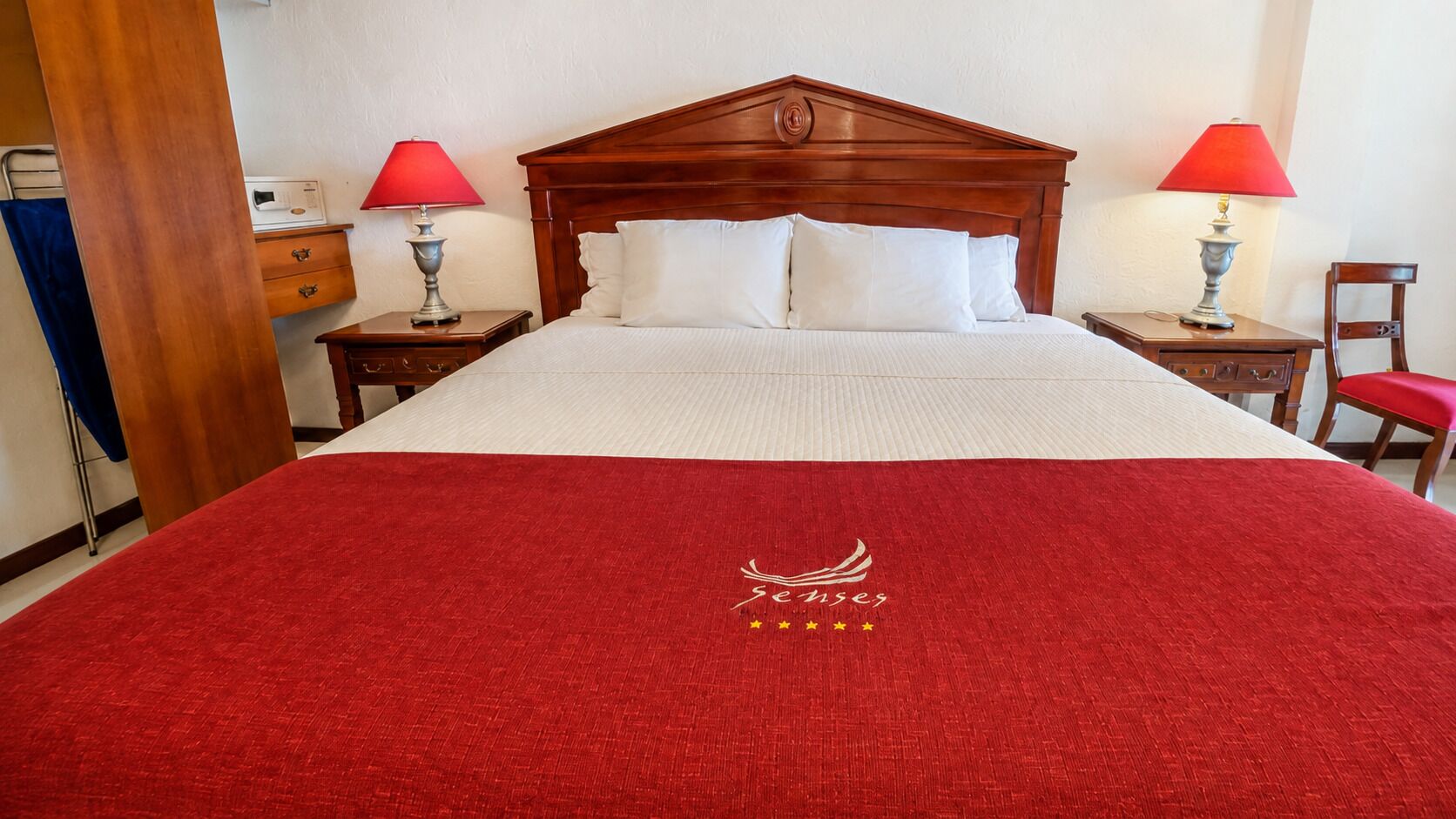 A hotel room with a large bed, two bedside tables with lamps, a wooden headboard, and a red carpet with a logo.