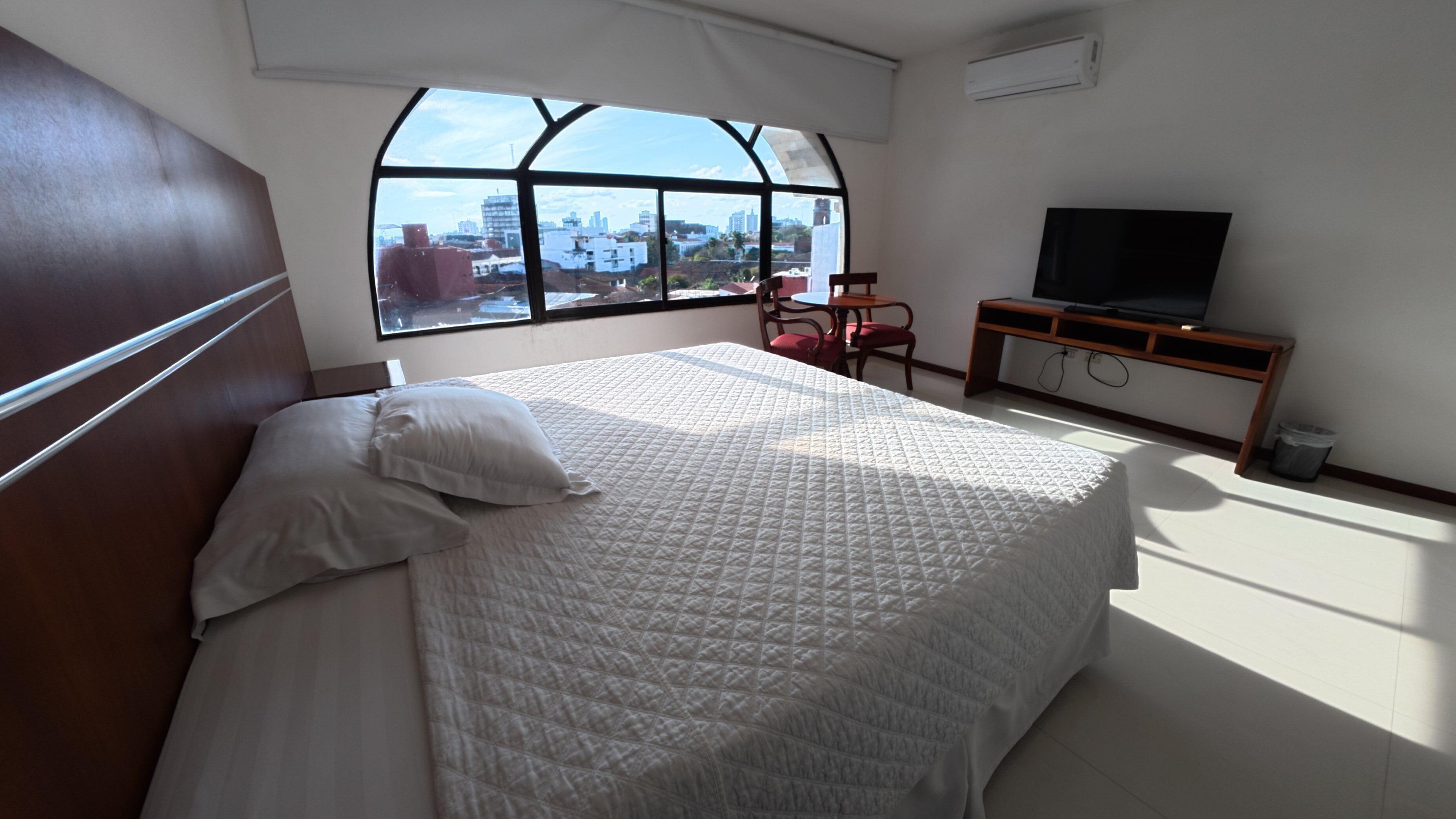 A hotel room with a large bed, a television, and a window with a city view.