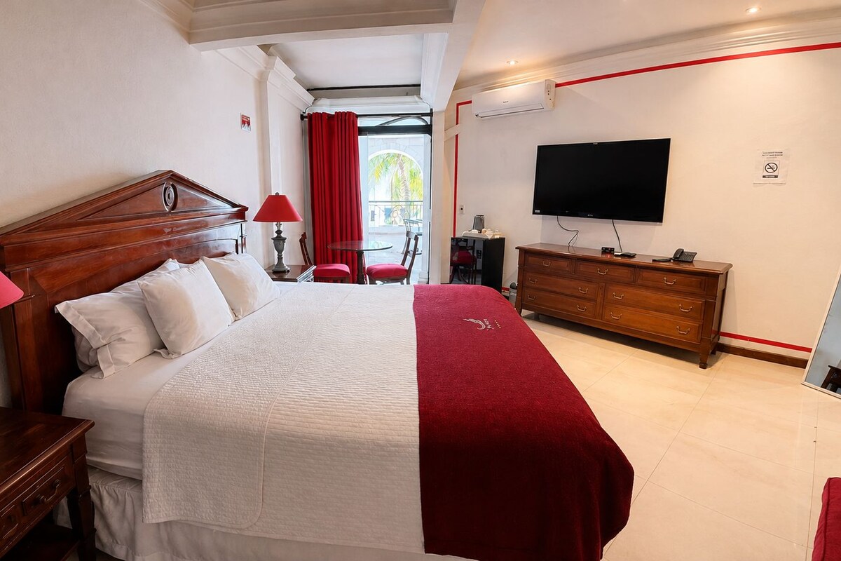 A hotel room with a large bed, a wooden dresser, a television, and a balcony with red curtains.