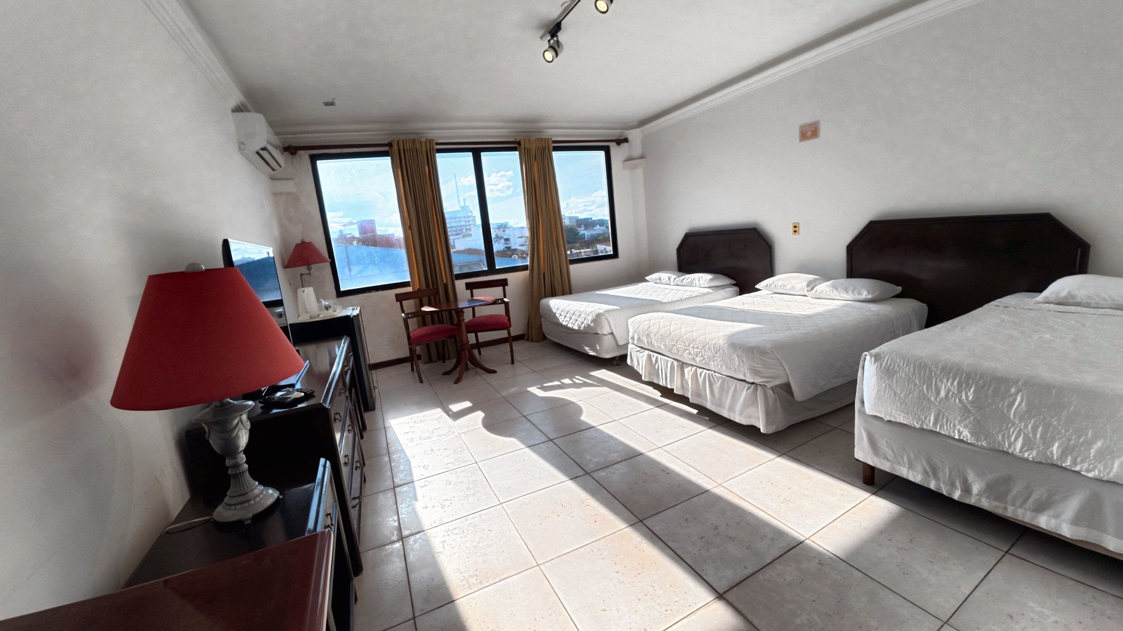 A hotel room with two beds, a desk with a lamp, a chair, and a view of the city through large windows.