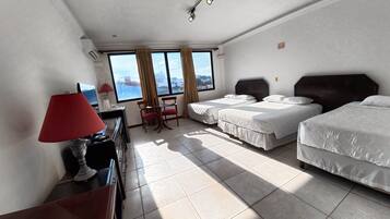 A hotel room with two beds, a desk with a lamp, a chair, and a view of the city through large windows.
