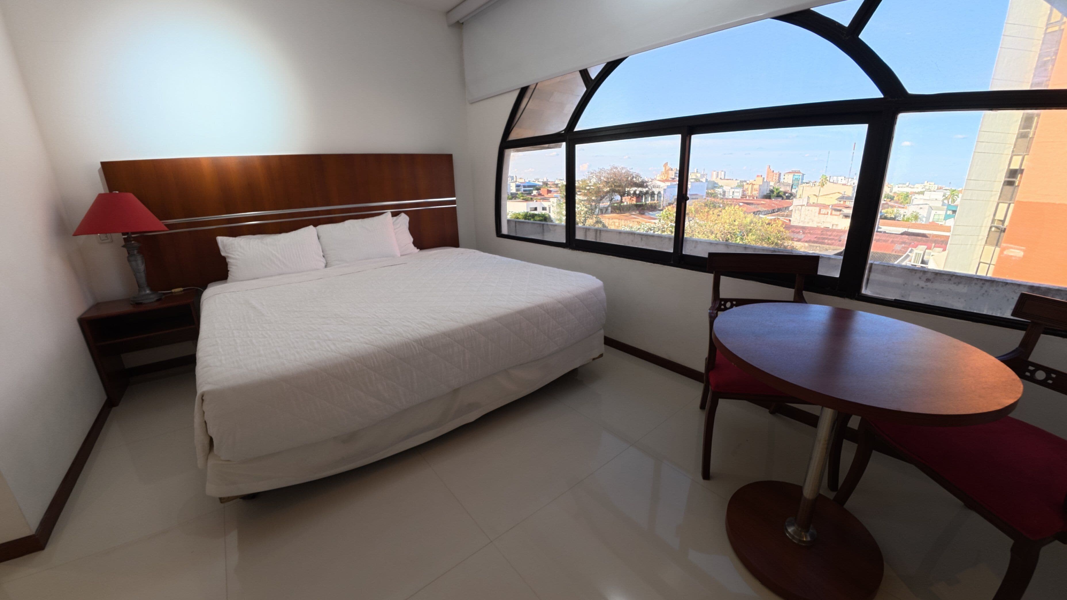 Quarto de hotel com cama, mesa de cabeceira, mesa e janela com vista para a cidade.