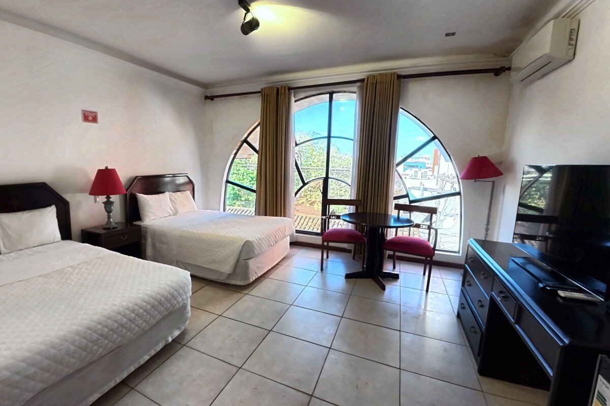 Habitación de hotel con dos camas, televisión, una mesita y balcón con vistas.