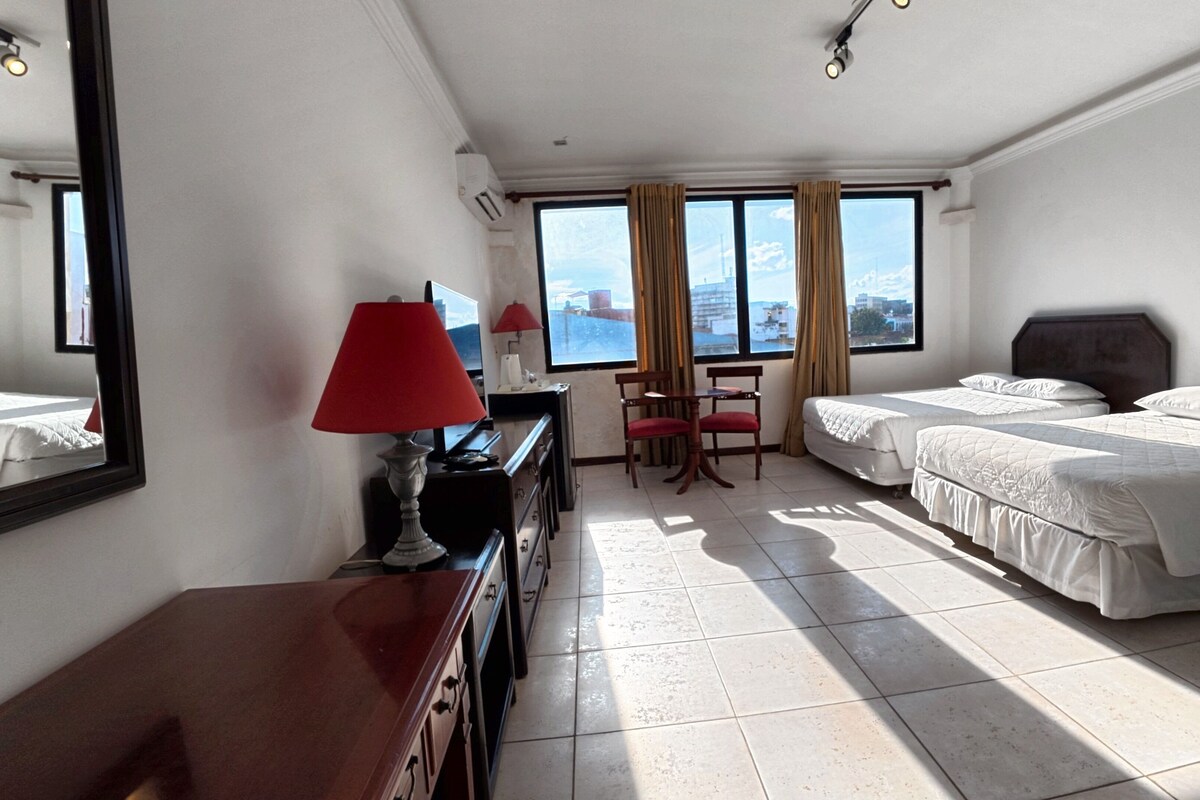 Una habitación de hotel con dos camas, un escritorio con lámpara, una silla y un ventanal con cortinas.