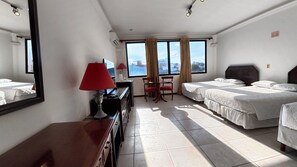 Una habitación de hotel con dos camas, un escritorio con lámpara, una silla y un ventanal con cortinas.