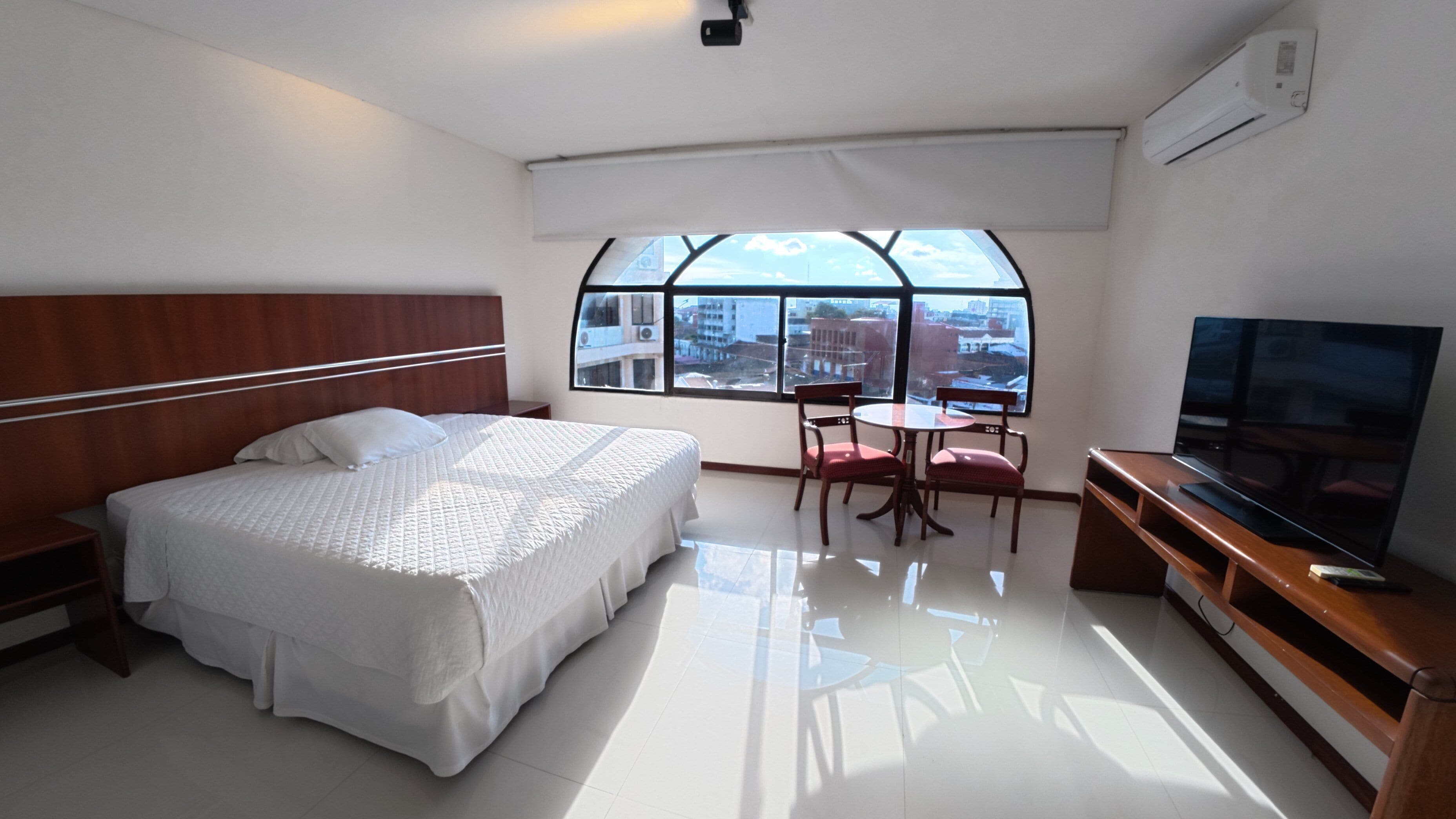 Quarto de hotel com cama, TV, escrivaninha e cadeira, além de uma janela grande.