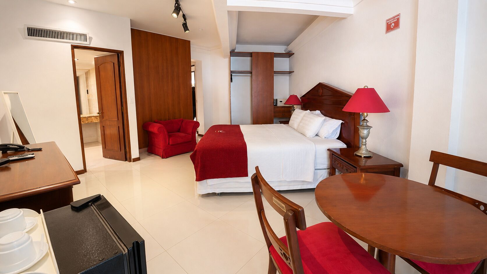 A hotel room with a bed, a desk, a chair, a table, and a red armchair.