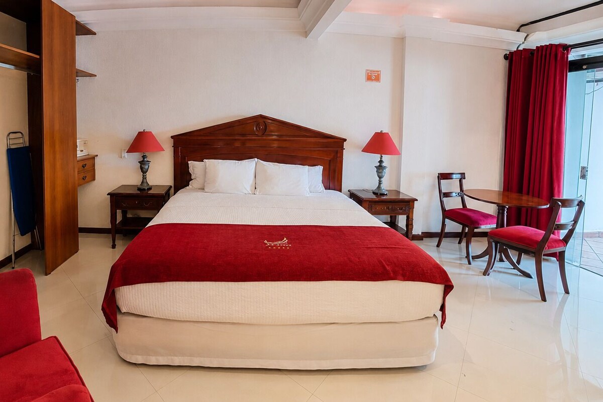 Una habitación de hotel con una cama grande, dos mesitas de noche con lámparas, una silla y una pequeña mesa con sillas.