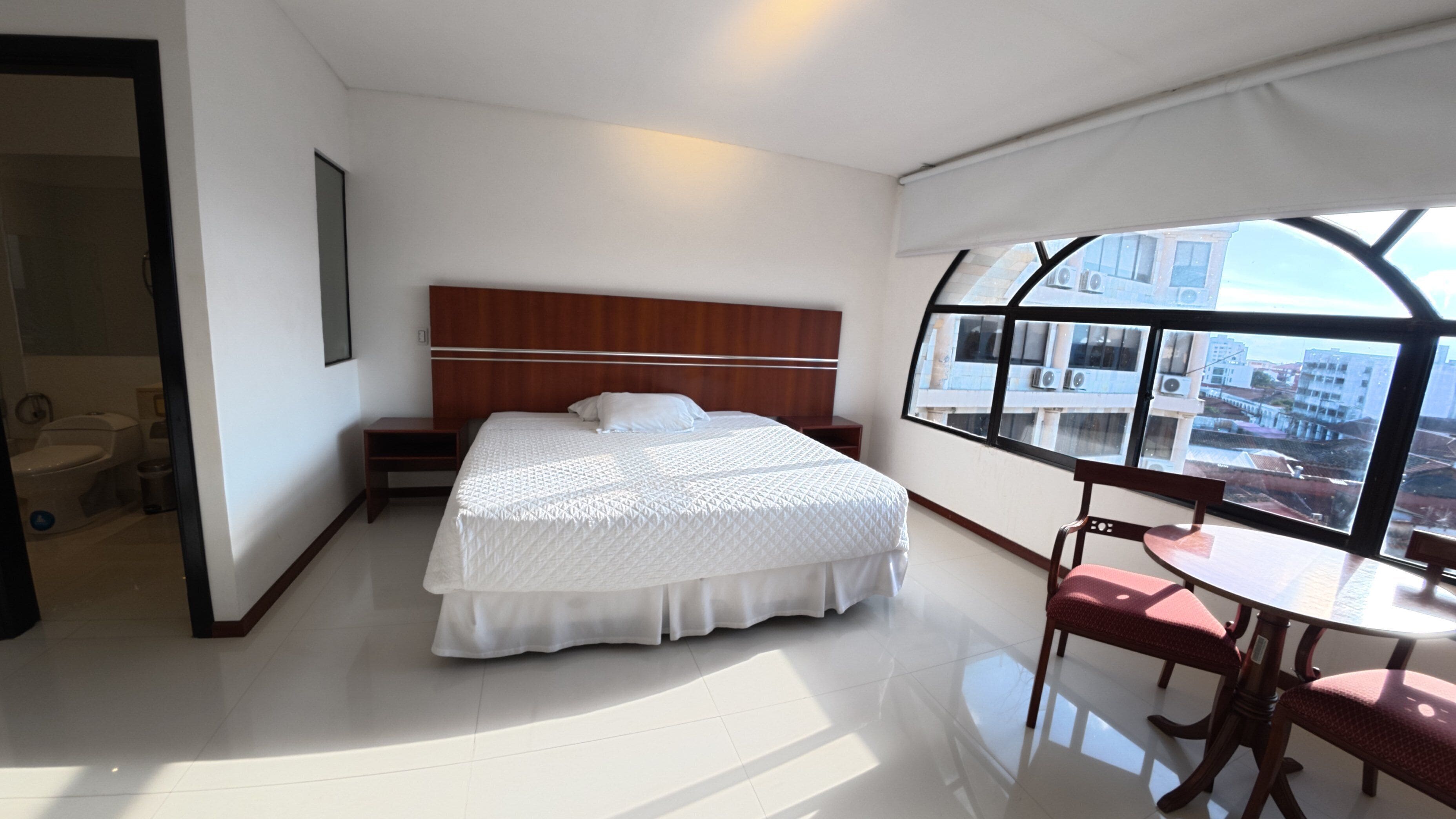 A hotel room with a large bed, a small table with chairs, a view of the city through a window, and a separate bathroom area.