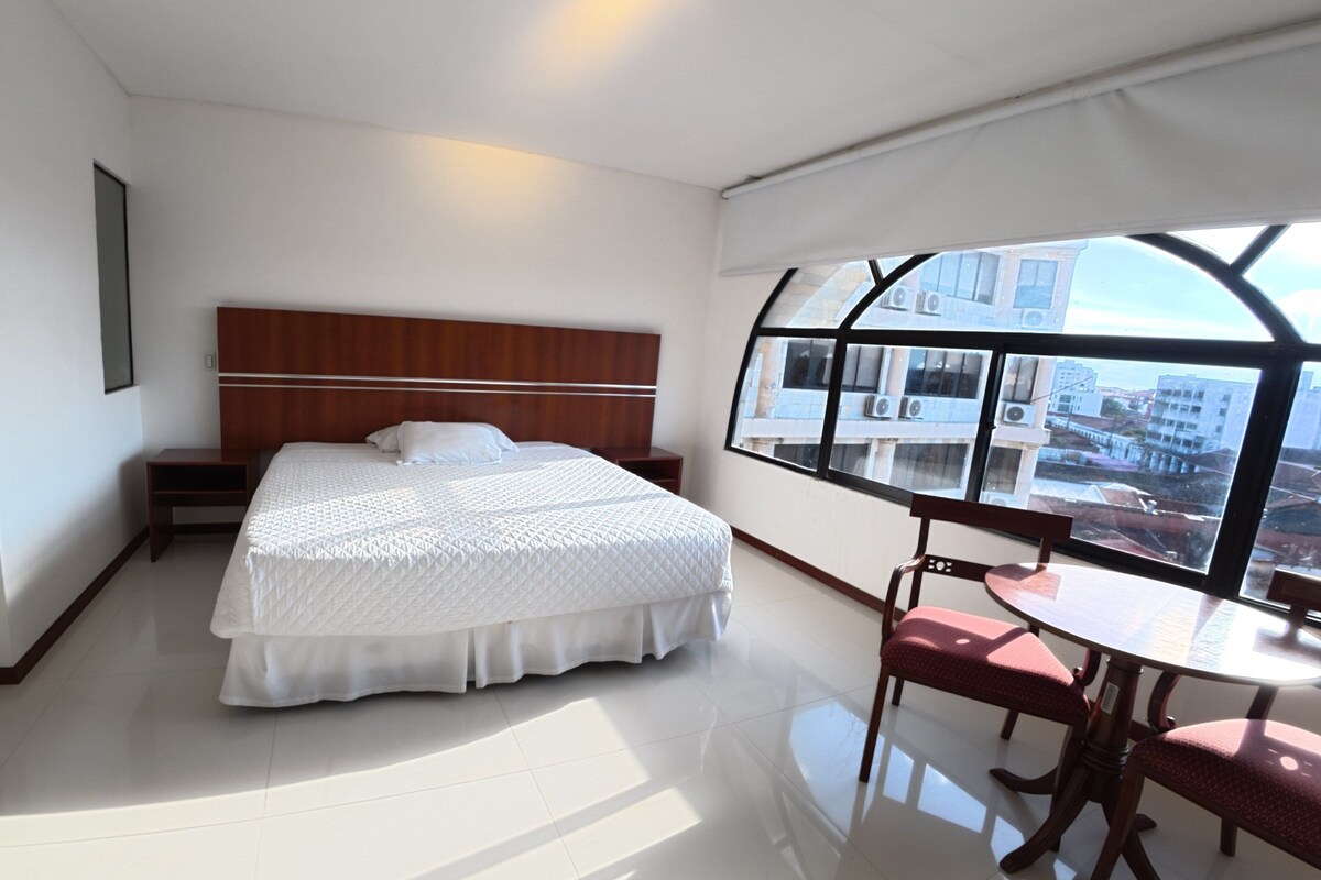Una habitación de hotel con una cama grande, una pequeña mesa con sillas, vistas de la ciudad a través de una ventana y un área de baño separada.