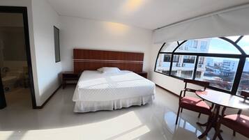 A hotel room with a large bed, a small table with chairs, a view of the city through a window, and a separate bathroom area.