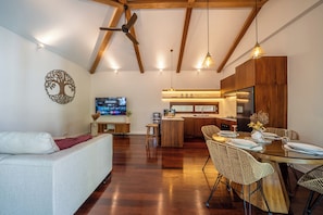 A modern living room with a sofa, dining area with a glass table, kitchen with wooden cabinets, and a ceiling fan.