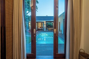 On aperçoit, à travers une porte vitrée, un espace piscine avec une terrasse en bois, une chaise longue et une table à manger.