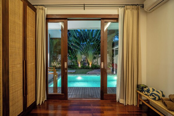 A room with a wooden floor, a sliding glass door leading to an outdoor pool area with a tropical garden view.