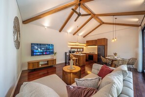 A modern living room with a high ceiling, a flat-screen TV, a sofa, and a dining area with wicker chairs.