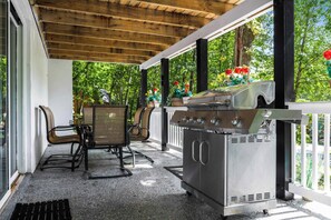 Een overdekt terras met een barbecue, zitplaatsen en uitzicht op bomen en bloemen.