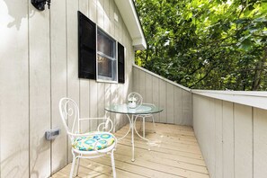 A patio with two white metal chairs and a glass table, a window with shutters, and a wooden deck.