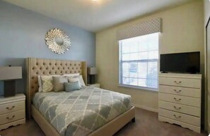 A bedroom with a tufted headboard, a bed with patterned bedding, bedside tables with lamps, a dresser with drawers, a television on top, and a window with blinds.
