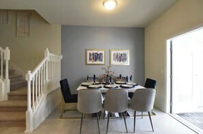 A dining area with a table set for four, a staircase, and two framed artworks on the wall.