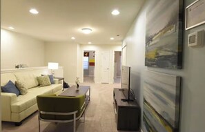 A living room with a sofa, a dining table, and a TV. There is a painting on the wall and a door leading to another room.