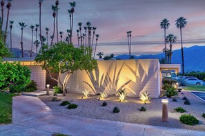 A modern building with a geometric facade, surrounded by a landscaped garden with cacti and palm trees, and a parking area with cars. - Seven Lakes Golf and Country Club 2 bedroom plus Den bungalow (Palm Springs)