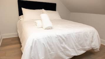 Cama bem arrumada com lençóis brancos e uma toalha enrolada, cabeceira escura e piso de madeira.