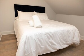 Cama bem arrumada com lençóis brancos e uma toalha enrolada, cabeceira escura e piso de madeira.