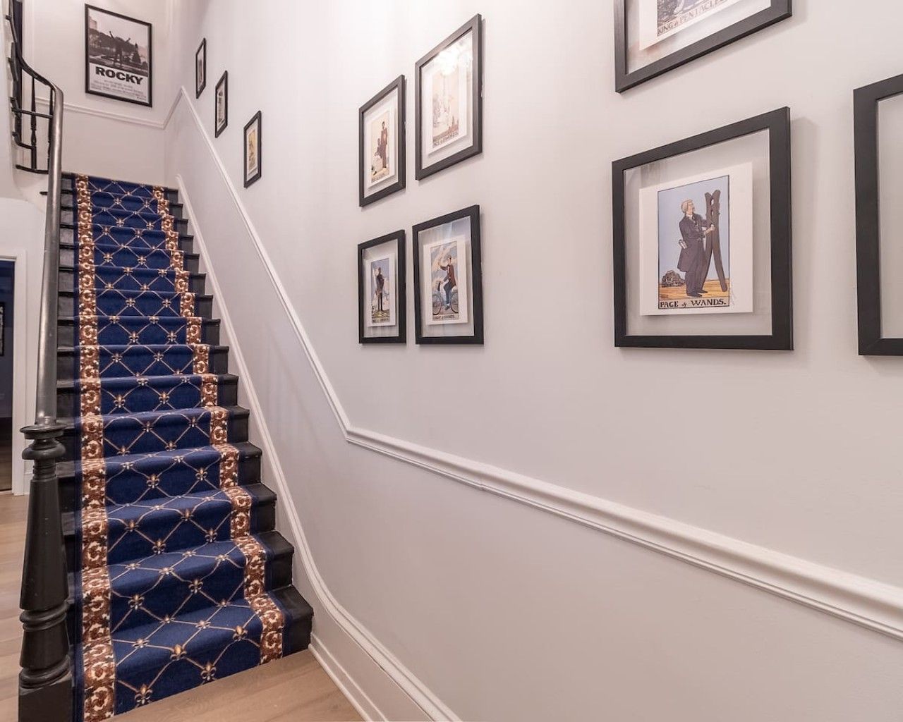Un escalier recouvert d’une moquette bleue et orné de dorures, menant à un palier où sont accrochées des images encadrées au mur.