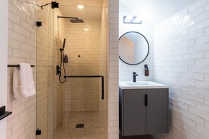 A modern bathroom with a glass-enclosed shower, a white-tiled wall, a round mirror, and a dark-toned cabinet. - Thena Hotel - Spacious Studio (Philadelphia)